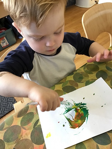 Preschool child sitting at the table holding a fork over some paint and the paper