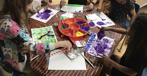 5 children sitting at a table with a tray of paint and multiple materials for painting