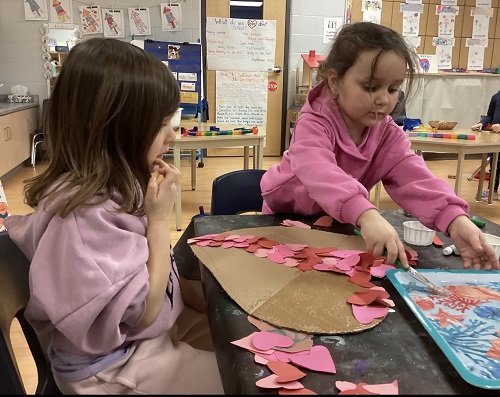 Two children gluing small paper hearts onto a large cardboard heart