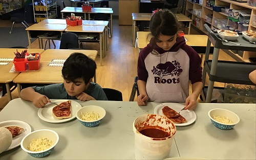 Two school aged children spreading pizza sauce onto the pizza crust using a spoon