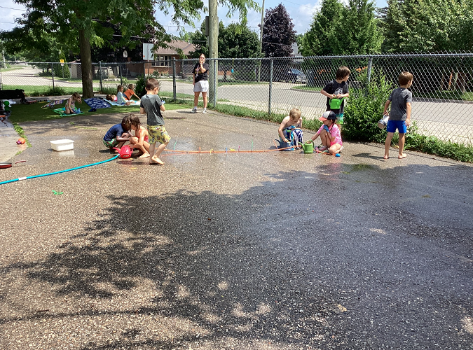 5 Summer camp children playing with the water sprinkler, 2 standing by the gate looking at the cars and 3 children sitting under the tree on their towels, while an educator observes