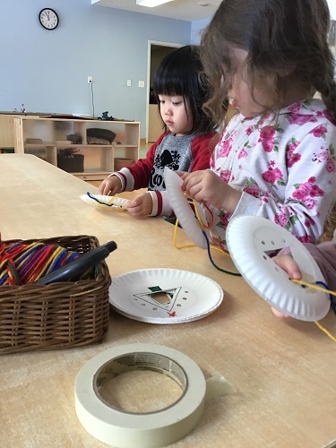 Two preschoolers are stringing yarn through a paper plate to make trees.