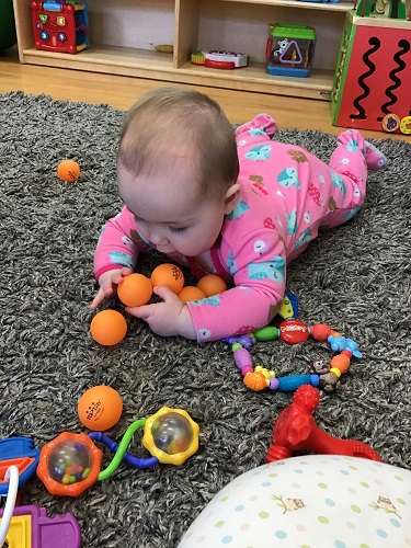 An infant is laying on their tummy with several ping-pong balls gathered in their arms.