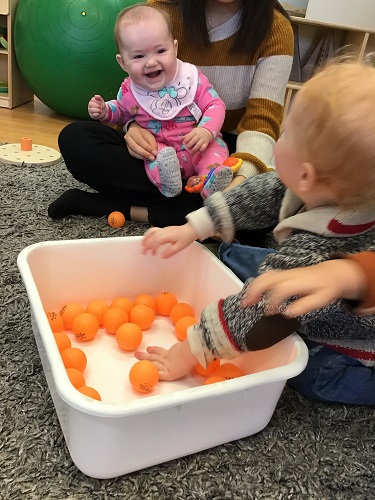 An infant is moving ping-pong balls around in a bucket as another infant watches.