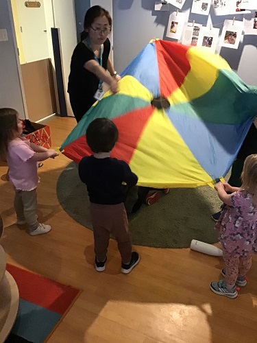 An educator and some preschoolers are playing a game with a parachute.