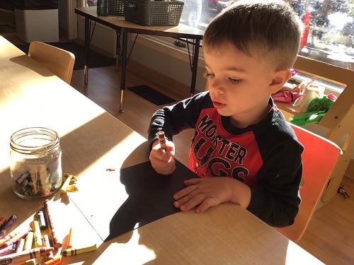 A preschooler is colouring with a crayon onto black paper.