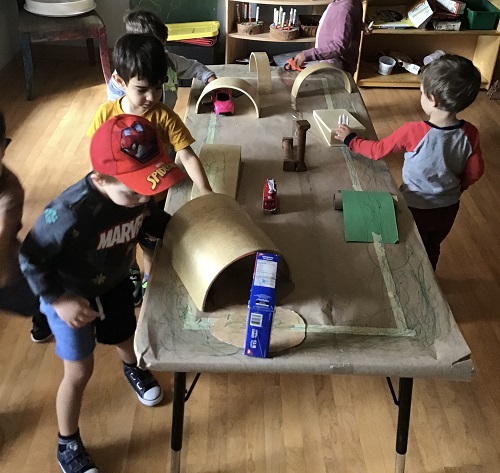 Several preschoolers are driving cars on a table top that they have created a road with tunnels.