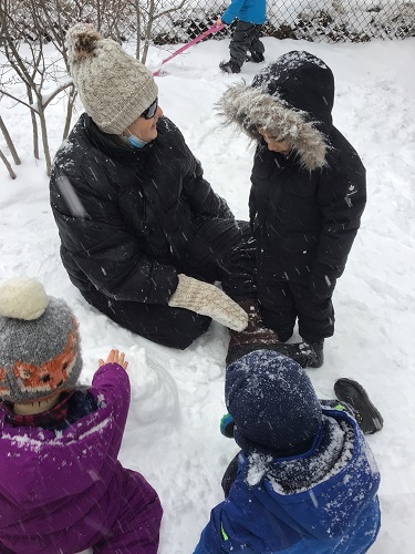 A preschool educator is with a group of children building a snowman.