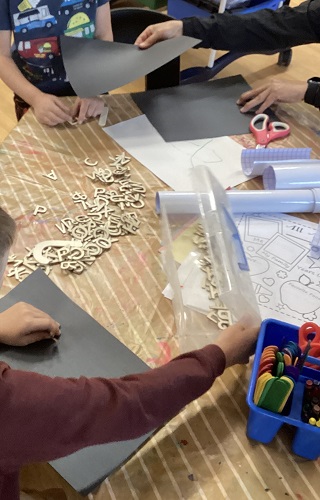 2 School Age 2 children with black paper and wooden letters and educator handing black paper to child