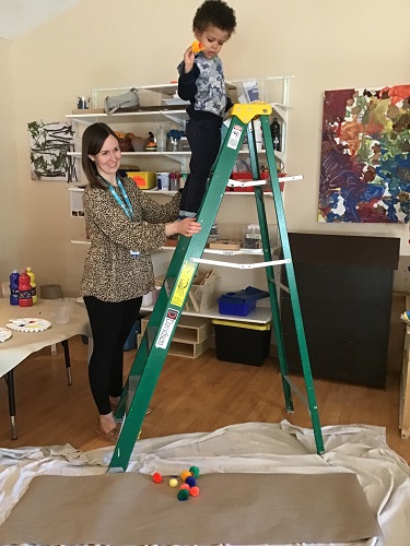 A preschooler is standing on a ladder, with an educator supporting them, and dropping their paint dipped pom pom onto paper.