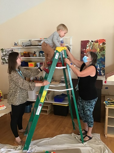 A preschooler is climbing to the top of a ladder, with an educator supporting them.