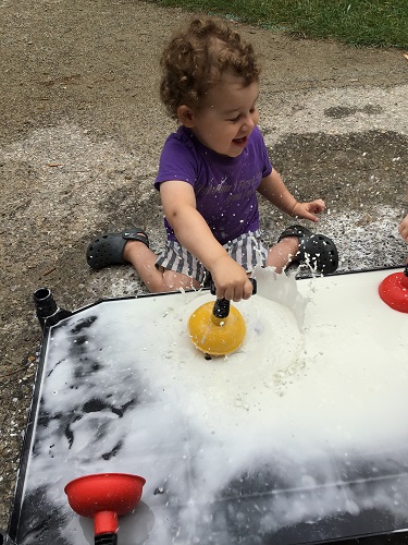 A toddler is using a small plunger to splash the Oobleck.