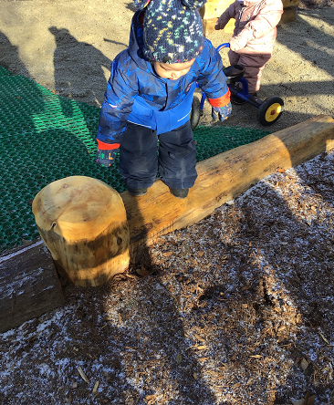 A toddler is about to jump off of a log that surrounds a play area on the new playground.