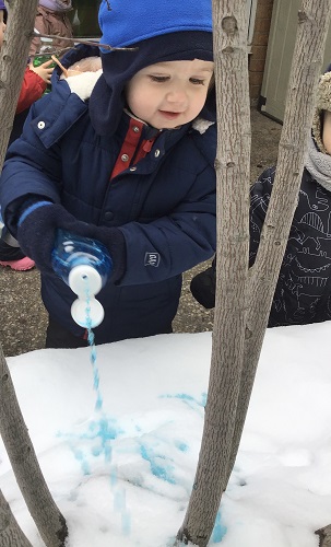 A toddler is squirting coloured water into some snow outside.