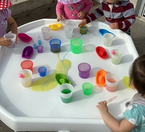 A group of children exploring with coloured water and various items in the white tuff tray.