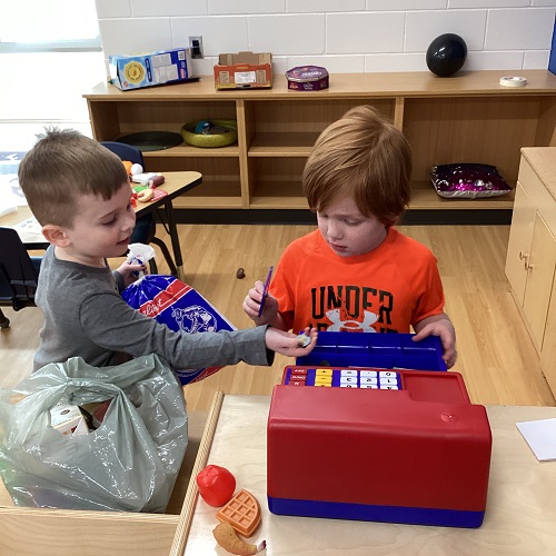 A child holding a food item and handing money to a child standing at a cash register.
