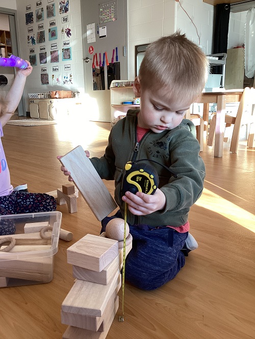 A child using a tape measurer to measure a stack of wood pieces.