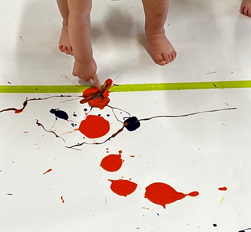 A child standing on a paper, dipping a finger in paint.