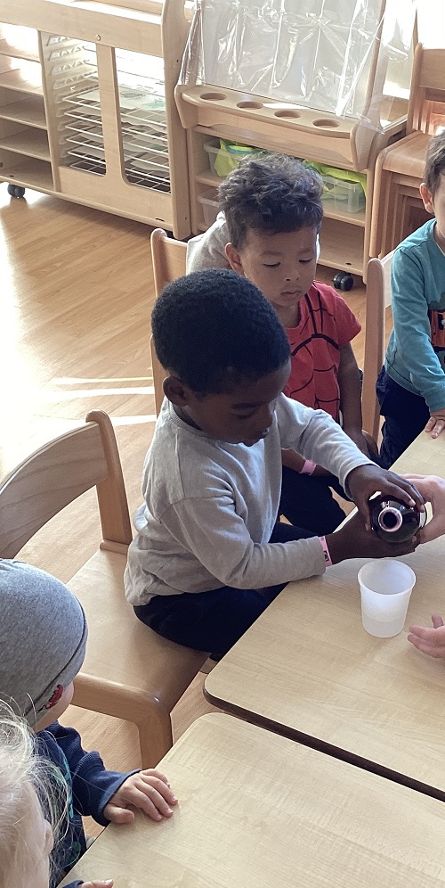 A child sitting down while pouring food colouring into a cup. A child beside them is observing what their peer is doing.