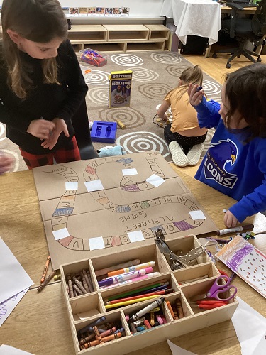 2 School Age 1 children standing at the table with the home made board game, looking at the board game and moving their pieces.