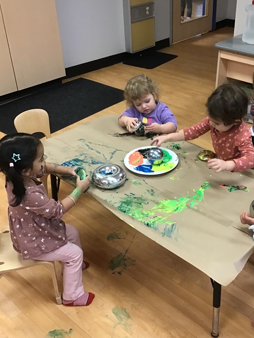 3 Toddler children sitting at the table with mirrored objects and paint, 1 toddler holding onto the mirror donut full of paint, another toddler dipping the mirror ball in the paint and another toddler sitting at the table with the mirror donut looking at her hands