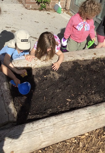 3 children crouching on the outside of the garden, one child touching the soil, one child using a spoon to pour water