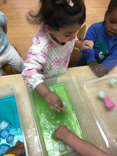 Toddler putting her hand in green water with ice cubes and another Toddler standing watching