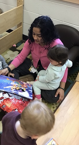 Toddler sitting in the teachers lap while she reads a story book