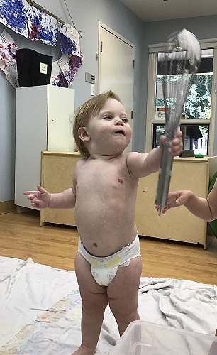 An infant is holding a whisk into the air that has bubbles on it.