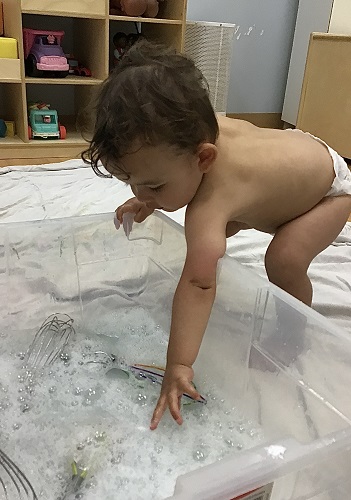 An infant is reaching into soapy water to grab a whisk.