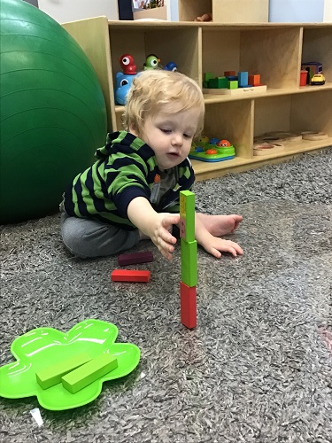 An infant is sitting in front of three coloured blocks stacked in a tower and is reaching out for the tower.