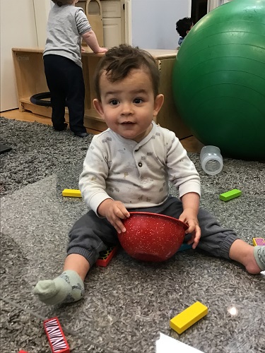 An infant is sitting on the floor with a bowl and surrounded by several coloured blocks.