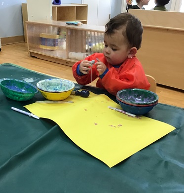 An infant has dipped their thumb into some coloured glue and is inspecting it closely.