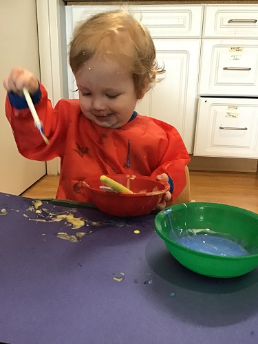 An infant is dripping coloured glue onto their paper.