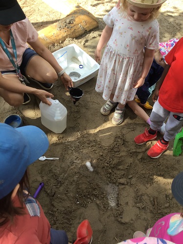 An educator, along with several preschoolers, are adding vinegar to their volcano.