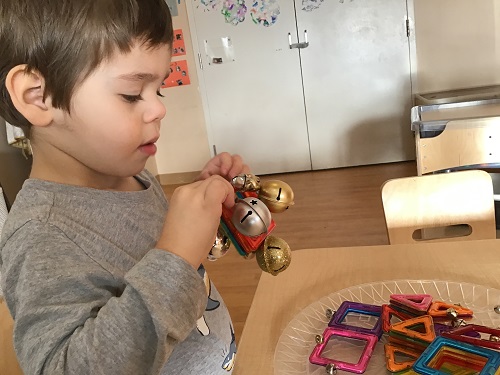 A preschooler is holding a magnetic tile block with metal jingle bells stuck to it.