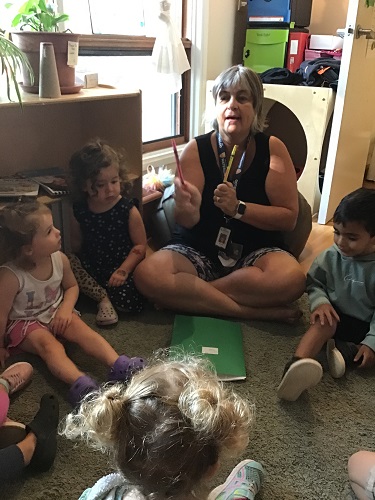 A preschooler educator is holding up two different coloured markers while sitting with a group of preschoolers.