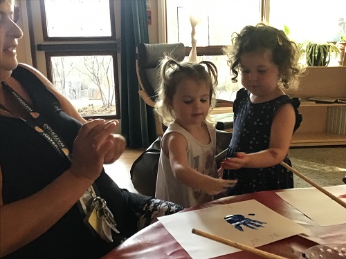 Two preschoolers are looking at their hands that have paint on them before making a hand print on paper.