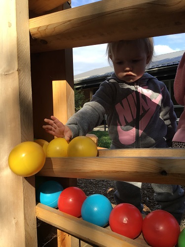 A toddler is pushing balls off the of the ball drop on the playground.