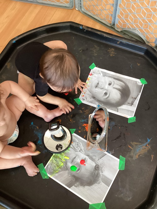 2 infants sitting on a tuff tray  with mirrors and photos of themselves and finger paint
