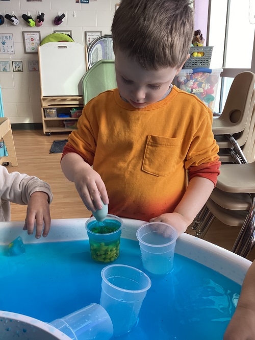 A preschool child holding a pipette in a cup filled with little ducks and blue water