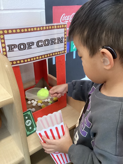 A preschool child scooping pom pom "popcorn" from their handmade popcorn machine into a container