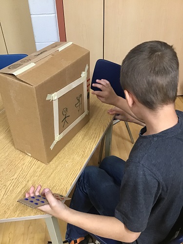 school age child with box on a table in front of them. Image of 2 stick figures boxed with tape and an imaginary  television remote in his hand 