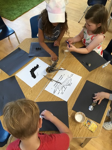  school age childre sitting at a table with black paper, glue , sticks and rocks and images of caterpillars 