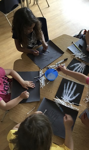 group of children  at a table with xrays of hands.  Each child  using brushes to add glue to the black paper before adding a white qtip