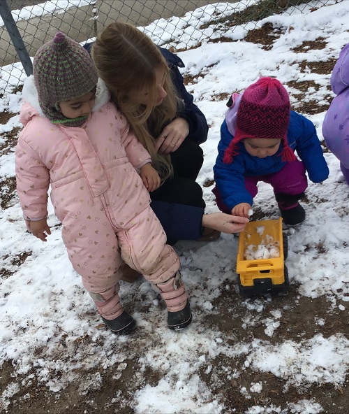 1 toddler pushing a truck in the snow, an educator and other toddler observing