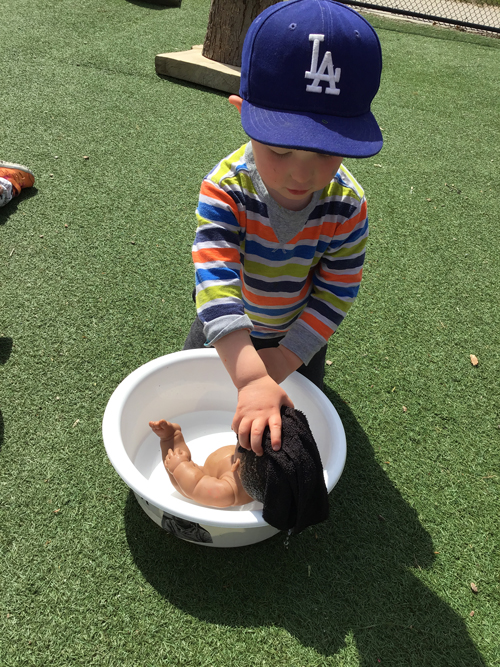 Boy bathing a baby doll in a round bin full of water outside