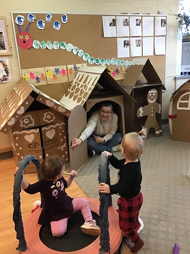 An educator sitting inside a big gingerbread house, inviting two children to come explore
