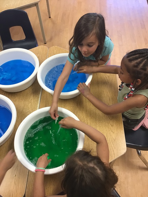 3 children sittnag at a table with 4 buckets 3 with blue water one with green water