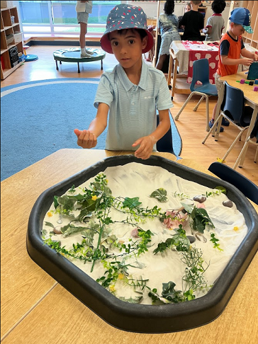 One Summer camp child standing with the tuff tray of sand and greenery holding his hand out and looking at the camera, there are 2 other Summer camp children standing away from the camera in the background